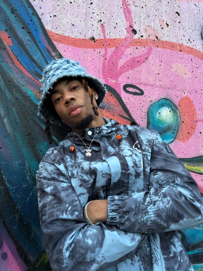 HUMBLE KD Young man in a stylish jacket and bucket hat posing against vibrant graffiti.